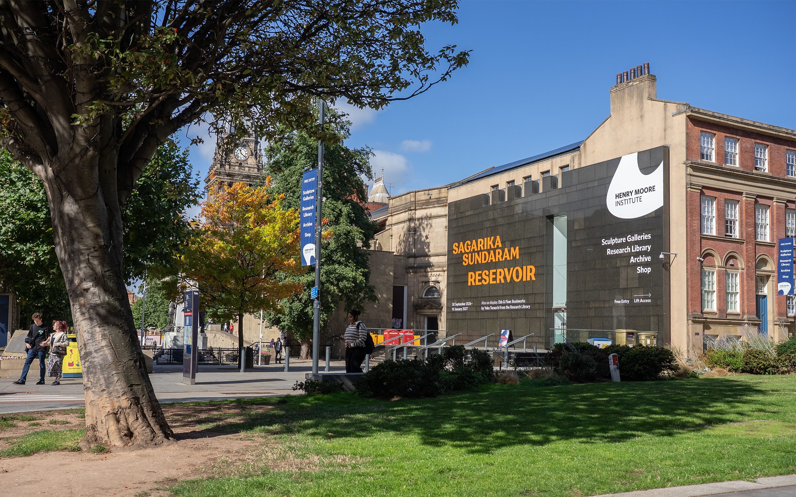 A three-story building with a black granite facade and steps leading up, with a leafy tree to the left. Vinyl lettering on the building reads: 'Sagarika Sundaram: Reservoir'.