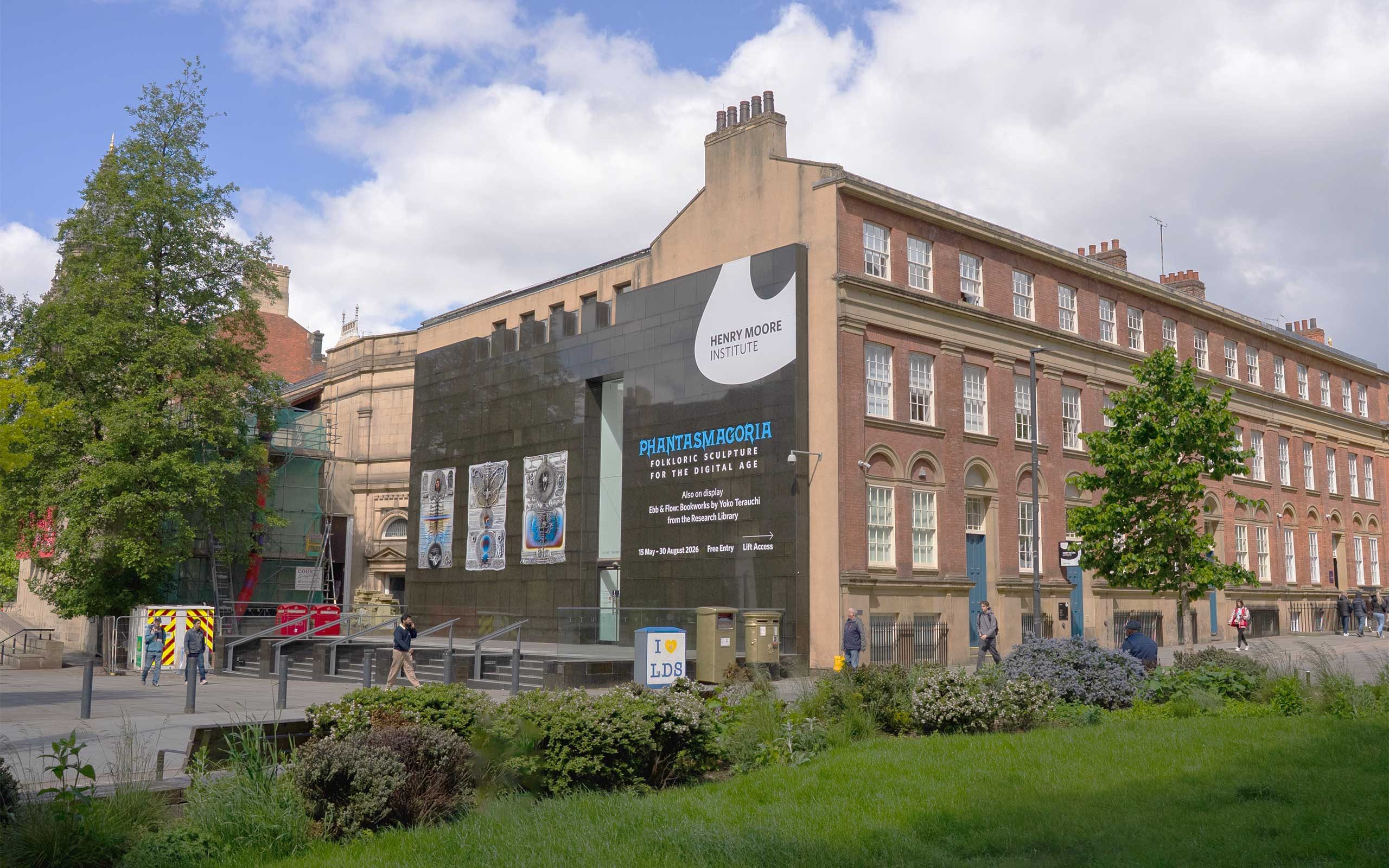 A three-story building with a black granite facade and steps leading up, with a leafy tree to the left. Vinyl lettering on the building reads: 'Phantasmagoria: Folkloric Sculpture for the Digital Age'.