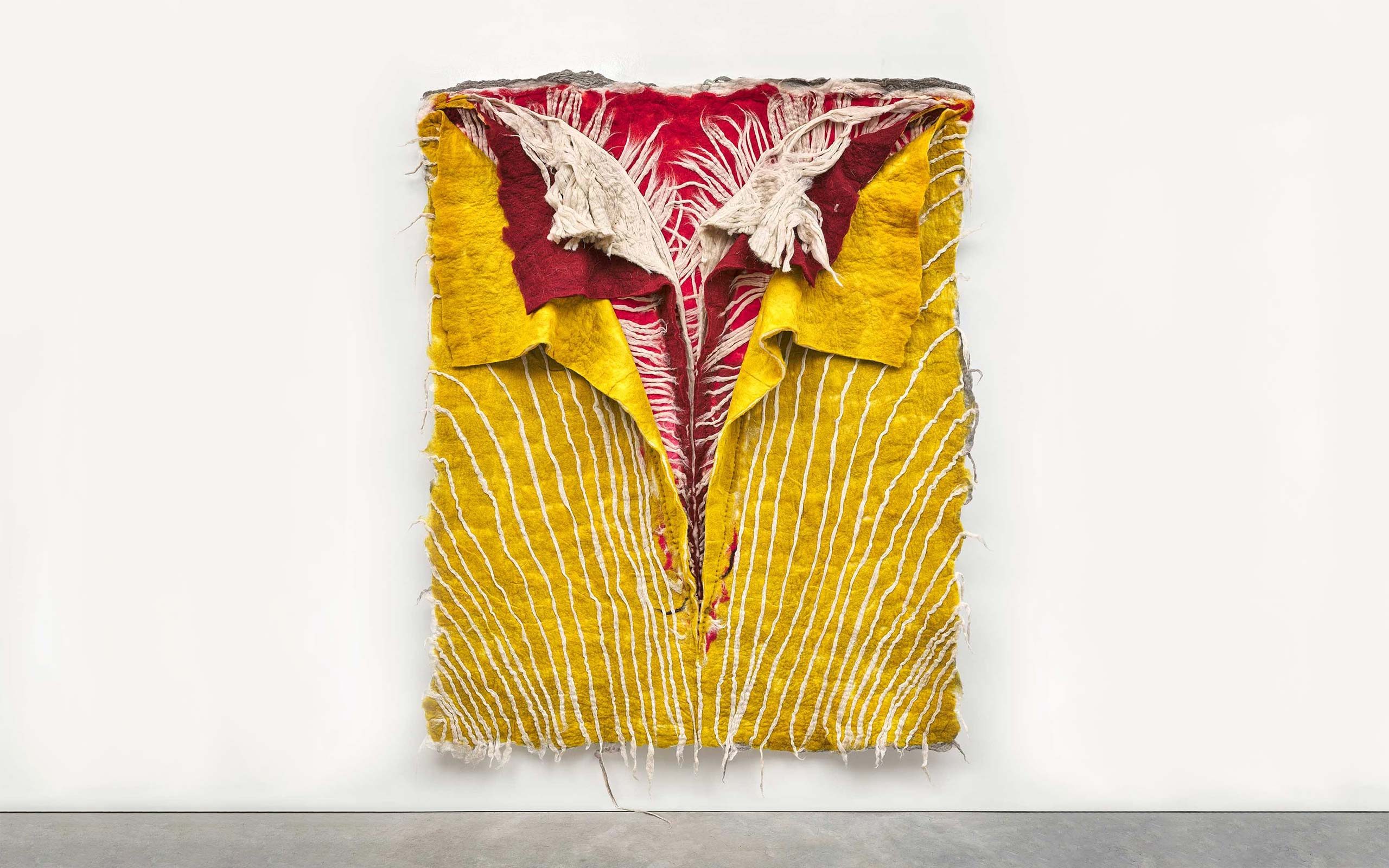 Artwork made of layered fabric, mounted on a white gallery wall. The topmost layer is yellow with white hand-stitched lines; underneath are layers of red and white fabric. They are opened and folded back at the top, similar to a layered shirt collar.