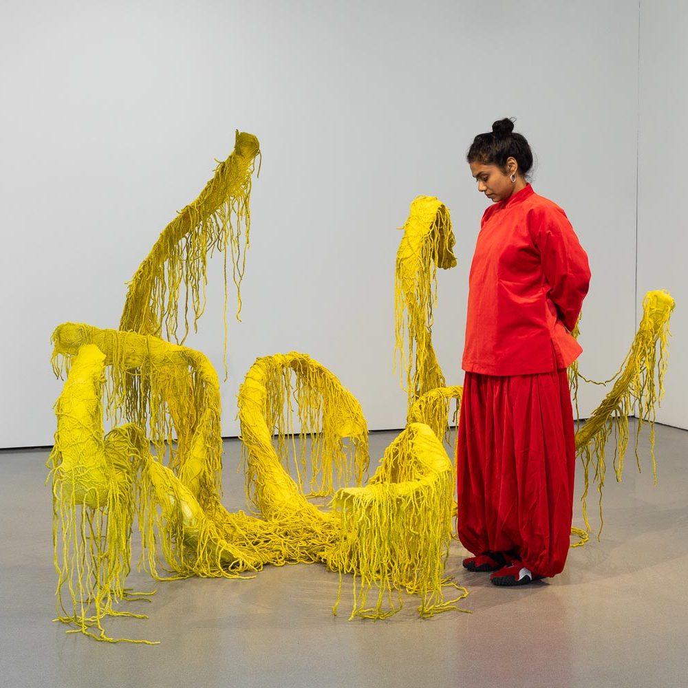 Artist Sagarika Sundaram standing next to her large fabric 'Glossolalia', which is made up of several yellow 'limbs', each trailing strands of yellow material.