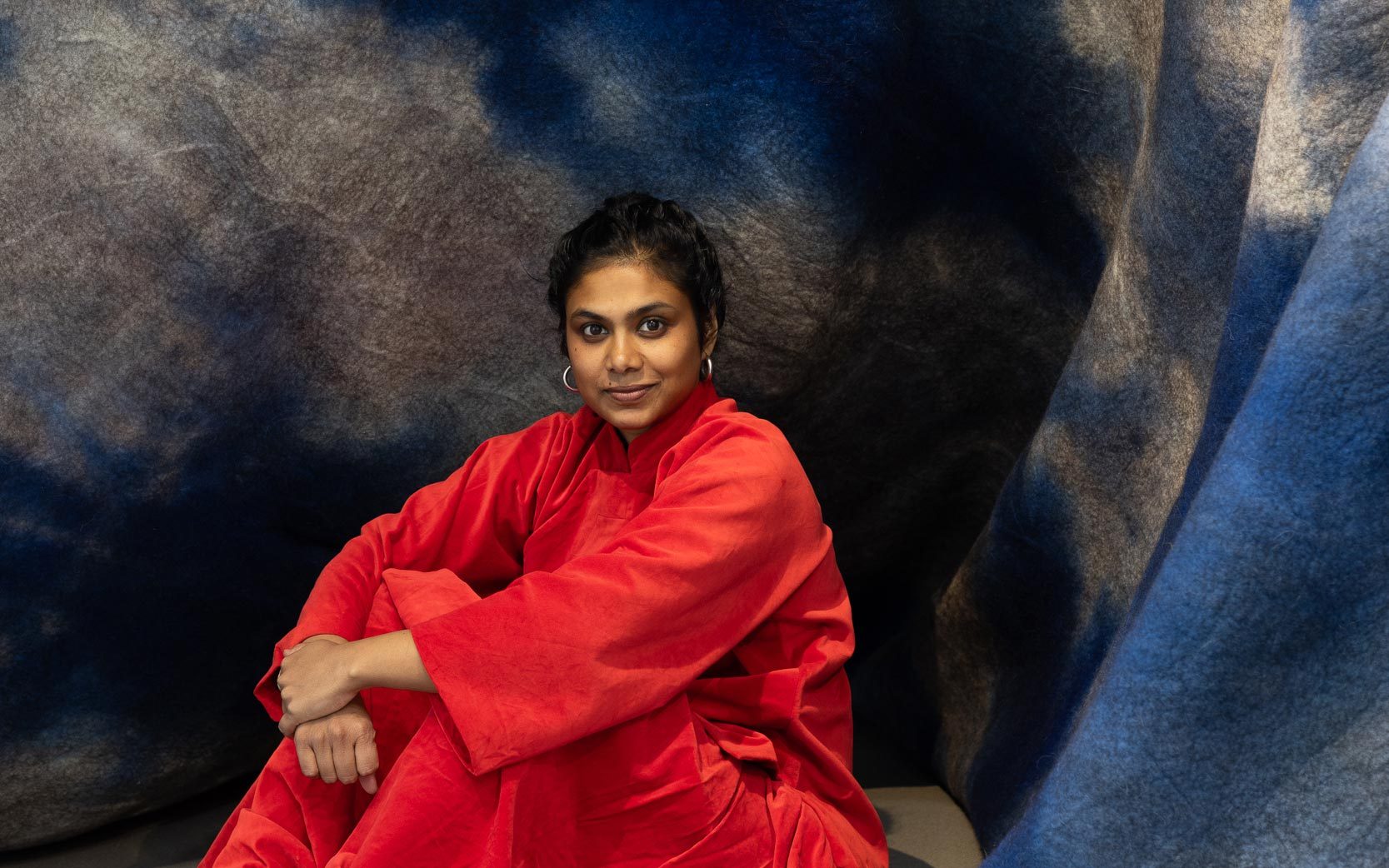 Artist Sagarika Sundaram sitting next to a large felt sculpture, which has been dyed blue and white and folded in a way that makes it look like a mountain or a wave crashing.