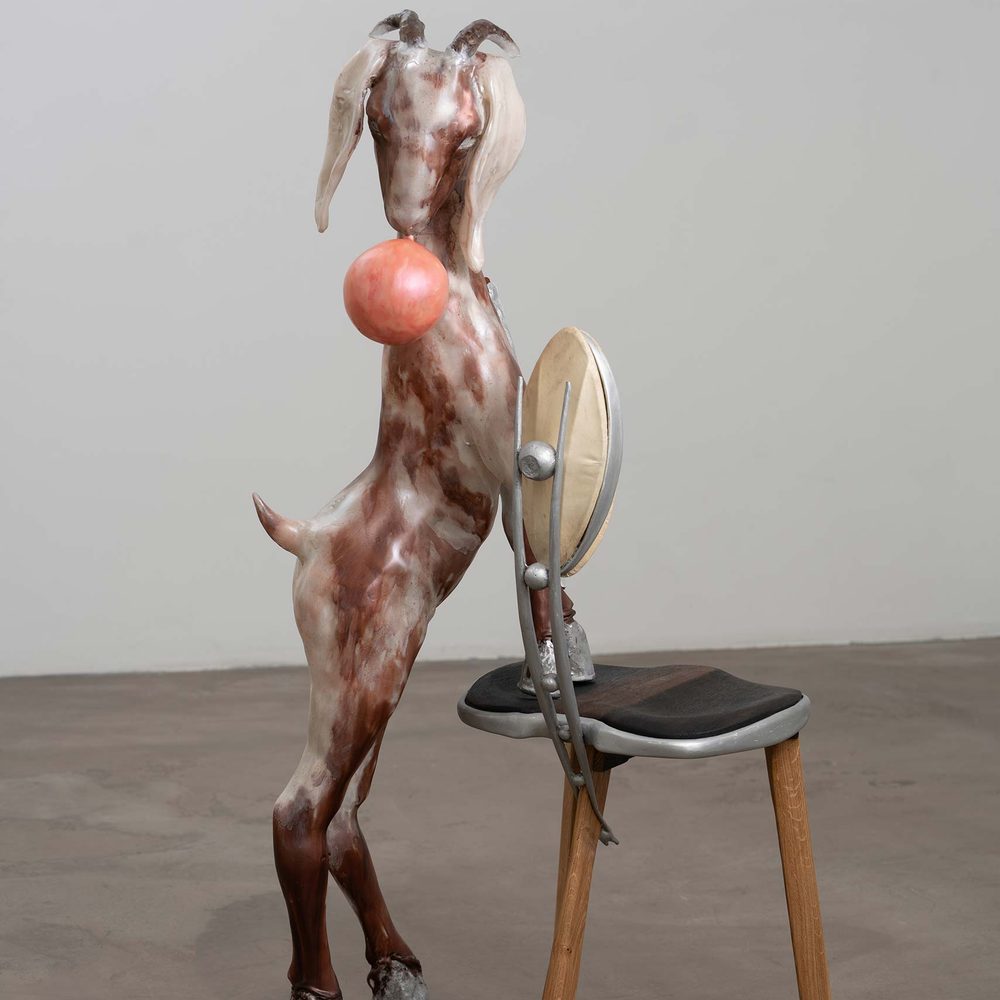 Contemporary sculpture of a goat-like creature with a ball held in its mouth, standing upright on its hind legs with its front legs resting on a chair.
