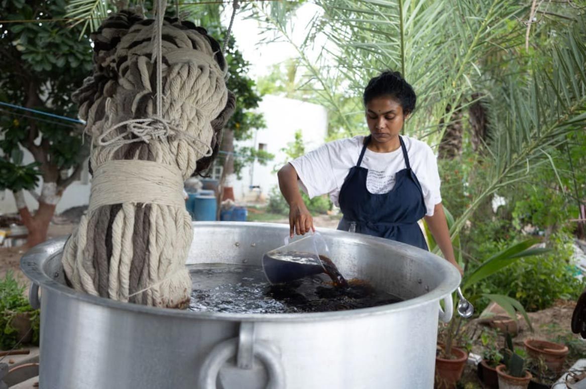 Artist Sagarika Sundaram stood outdoors, stirring a large pot of dye, into which a large piece of sculptural rope is being dipped.