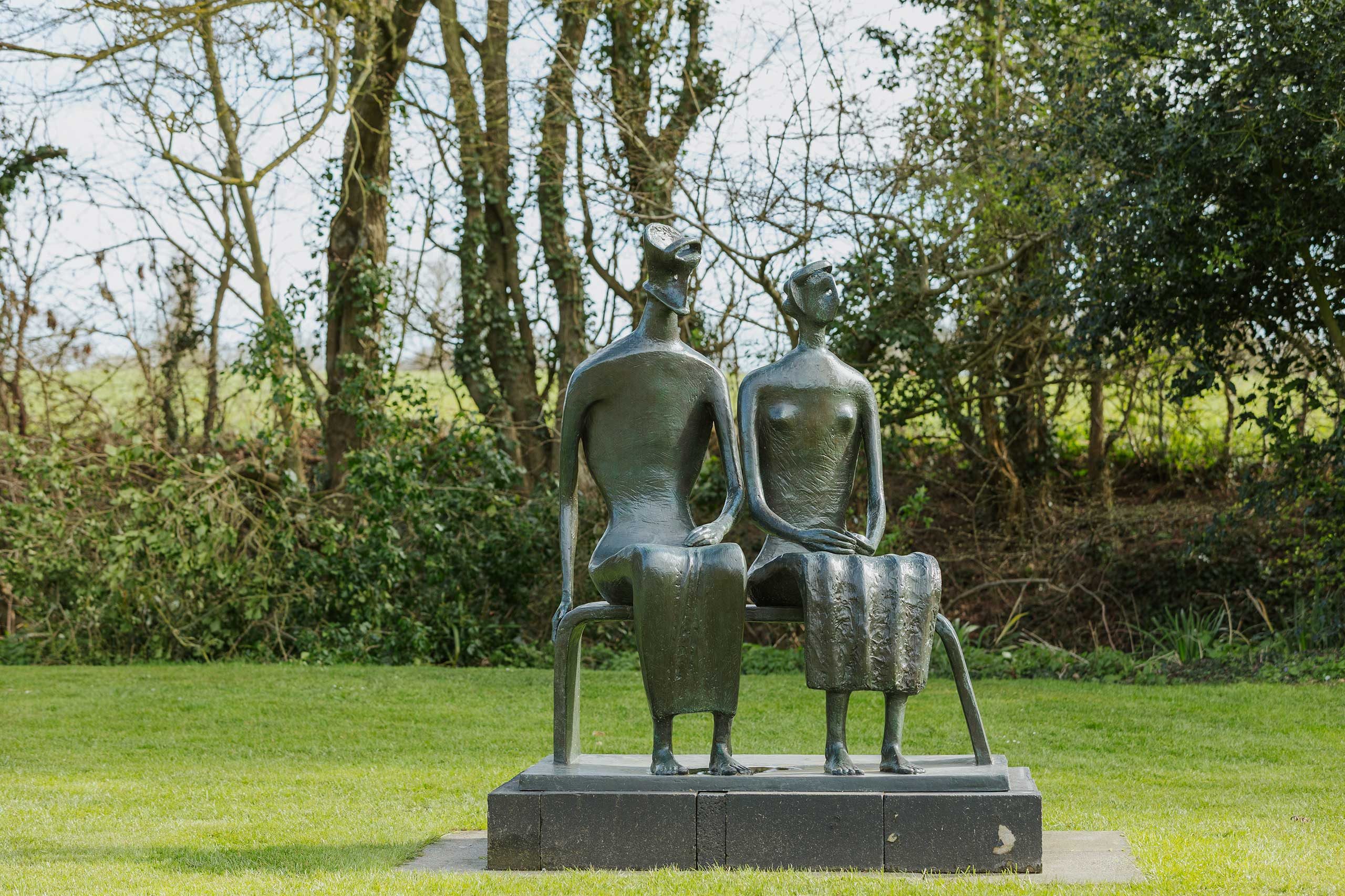 Bronze sculpture of two abstract figures sitting side by side on a bench, in a grassy field with trees behind.