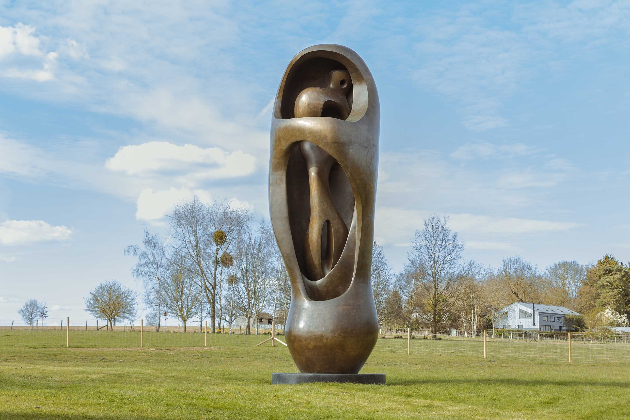 A large, abstract bronze sculpture of a person standing in a protective shell or cocoon. It is displayed on a plinth in a grassy field, with trees behind in the distance.