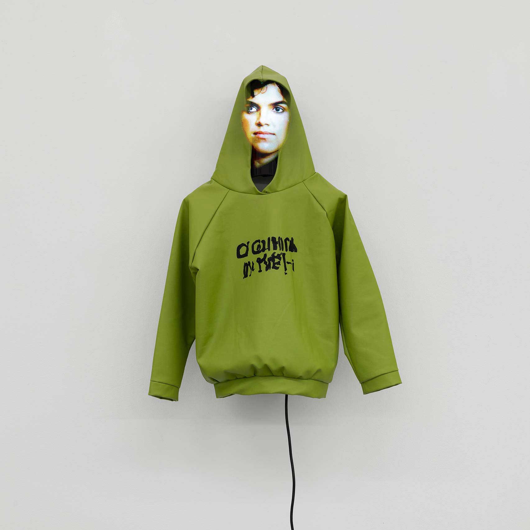 A green hoodie mounted on a white wall, with an illegible AI-generated slogan and a digitally generated 'face' under the hood. A wire trails down the wall from the hoodie's waist.
