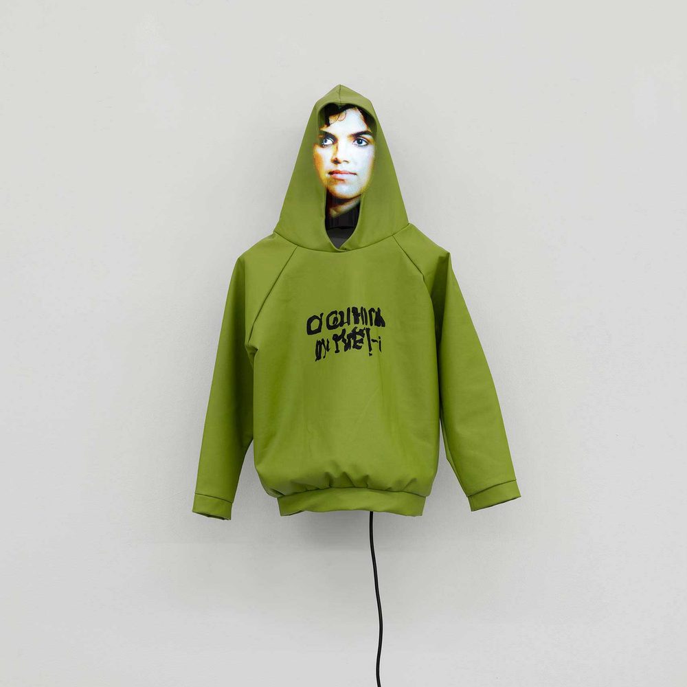 A green hoodie mounted on a white wall, with an illegible AI-generated slogan and a digitally generated 'face' under the hood. A wire trails down the wall from the hoodie's waist.