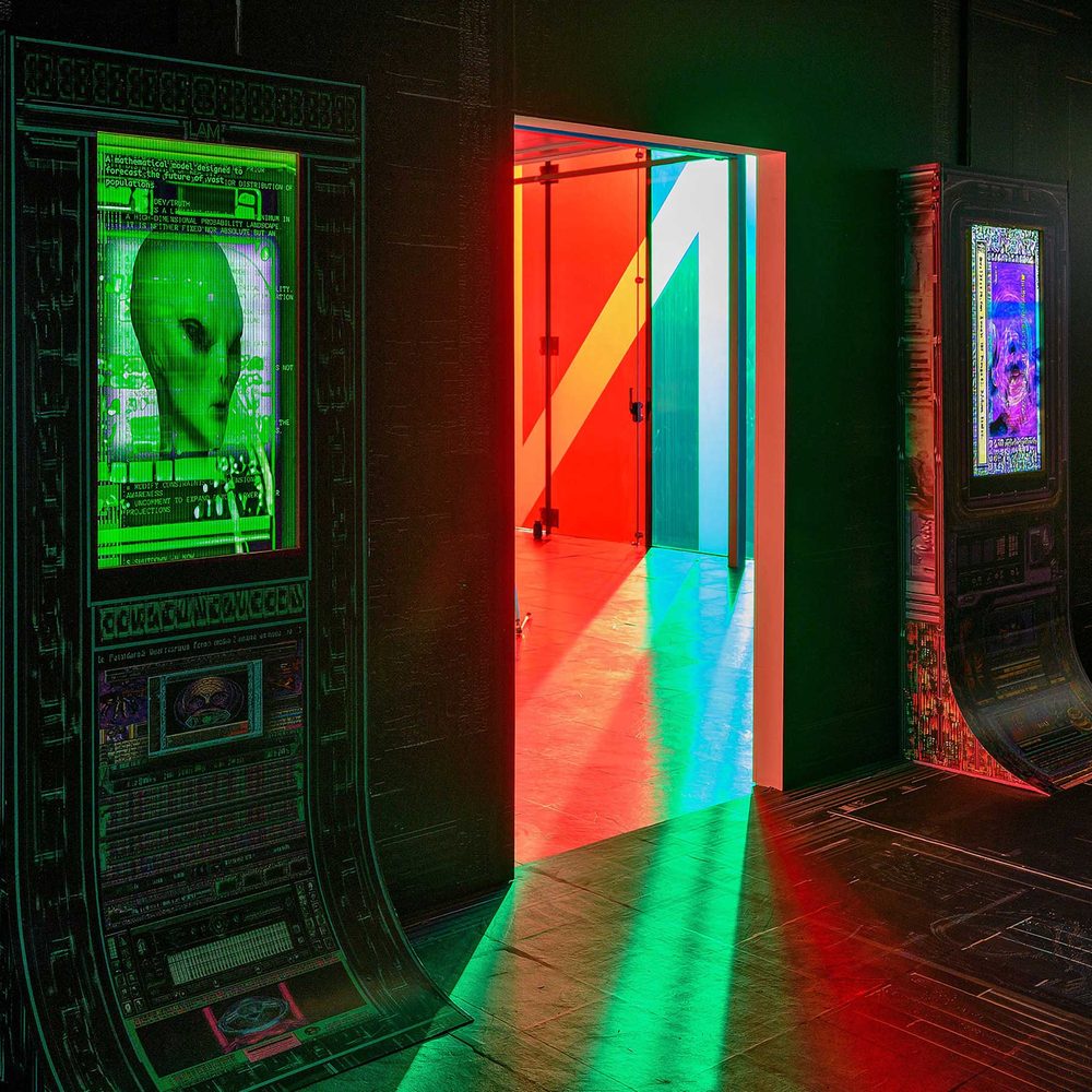 A futuristic or alien room, featuring wall-mounted display screens showing images of human and alien faces. The walls and other surfaces look to be made of a dark metal, lit by neon greens and reds.