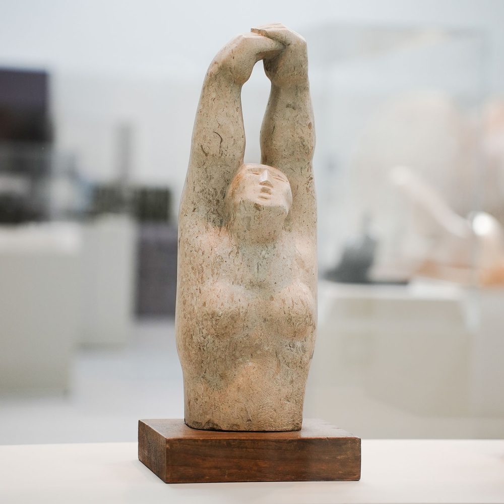 A limestone sculpture of a woman's torso, with her hands raised over her head.
