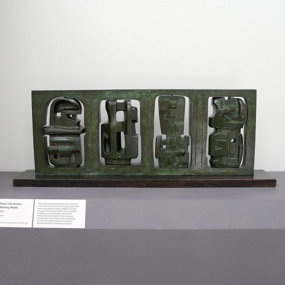 A long and flat, rectangular bronze sculpture by Henry Moore. It features four cut-out sections with intricate, figure-like sculptures inside.