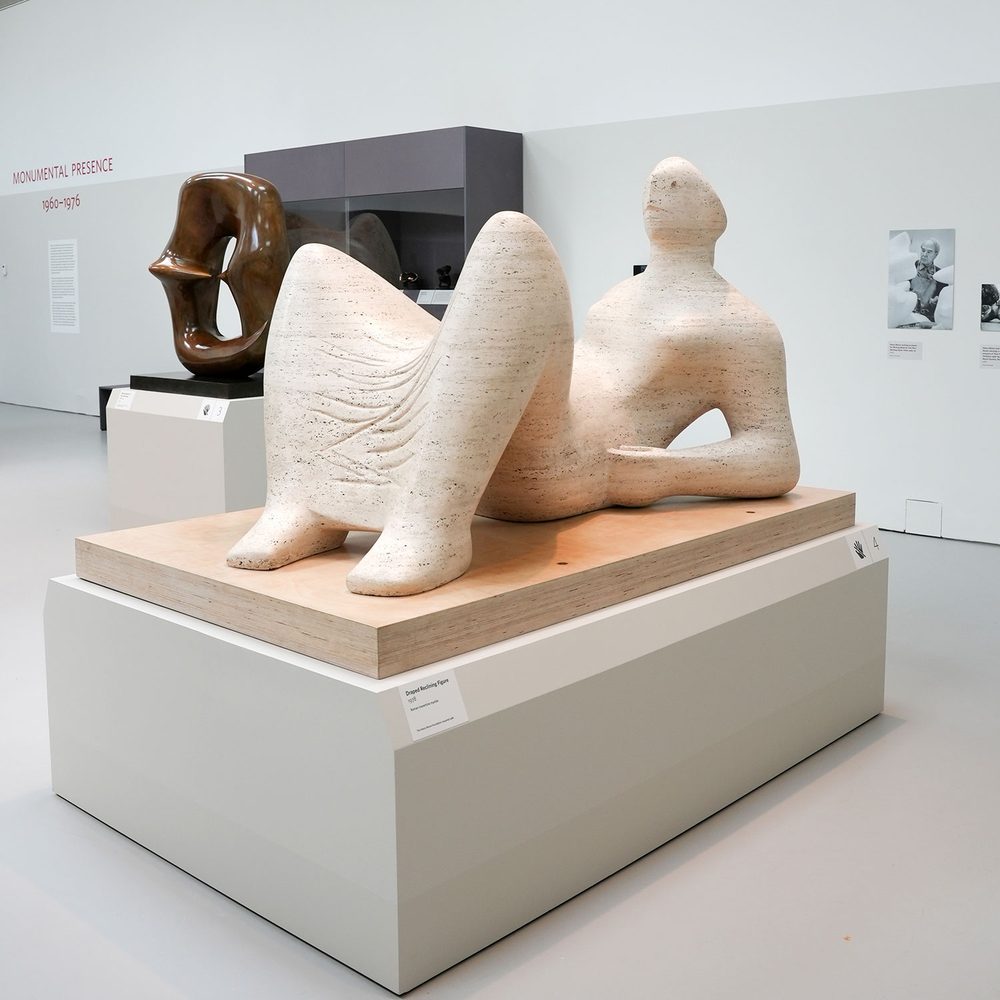 An abstract figurative sculpture or a reclining woman, carved from white marble. It is presented on a plinth in a white-walled gallery space.