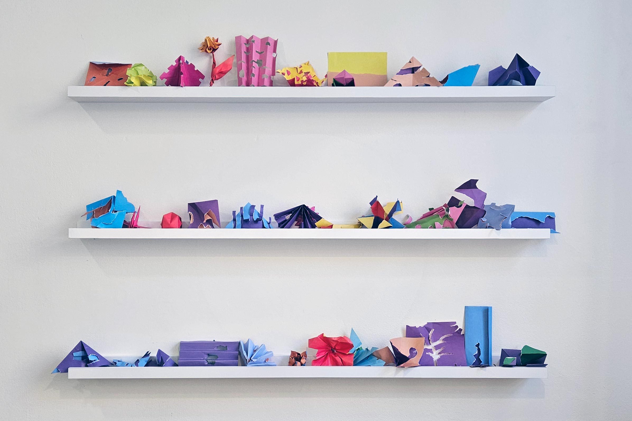 Three shelves displaying colourful sculptures made from torn and folded paper.