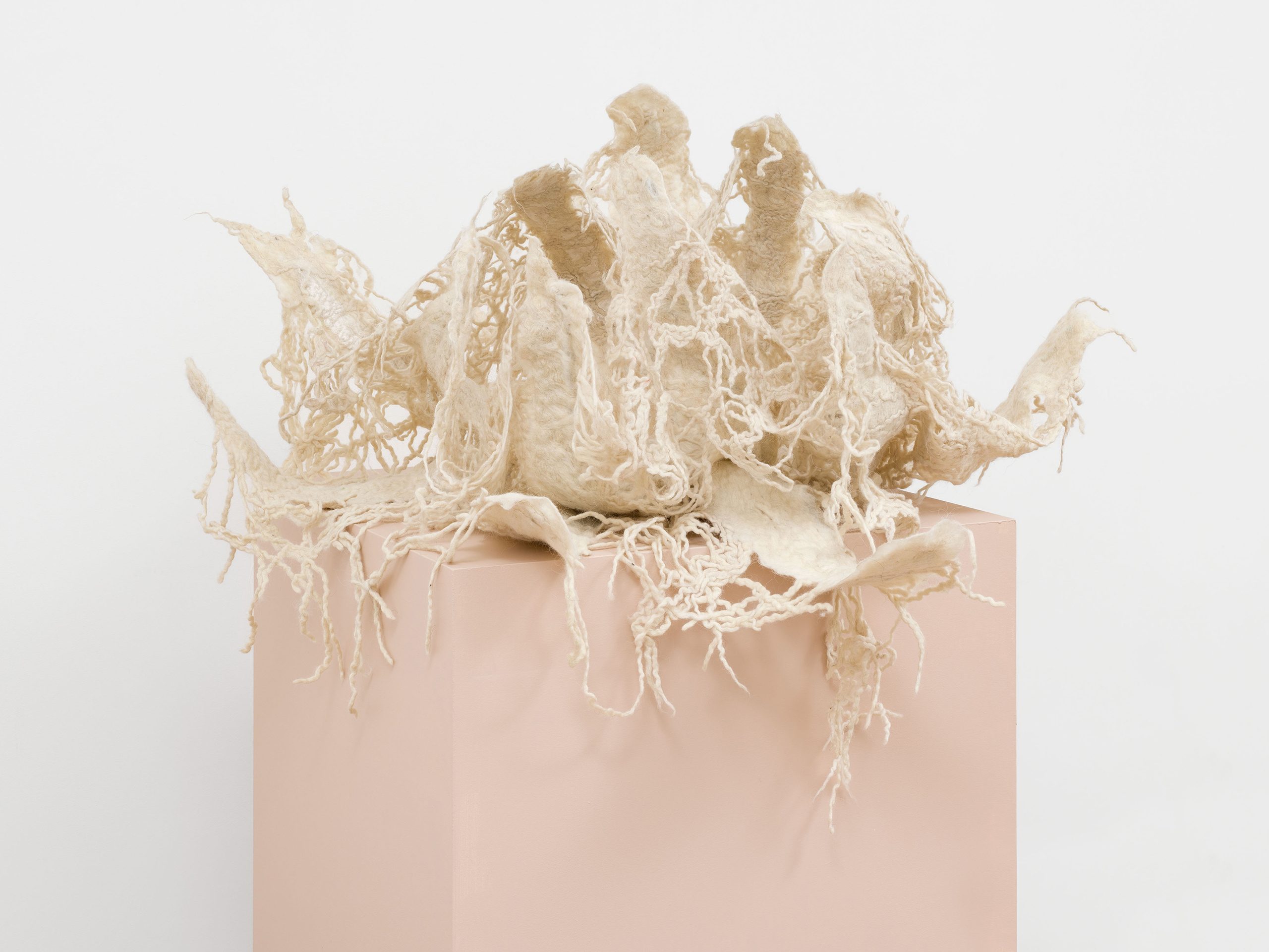 An abstract sculpture made of white wool and wire on top of a pale pink plinth.