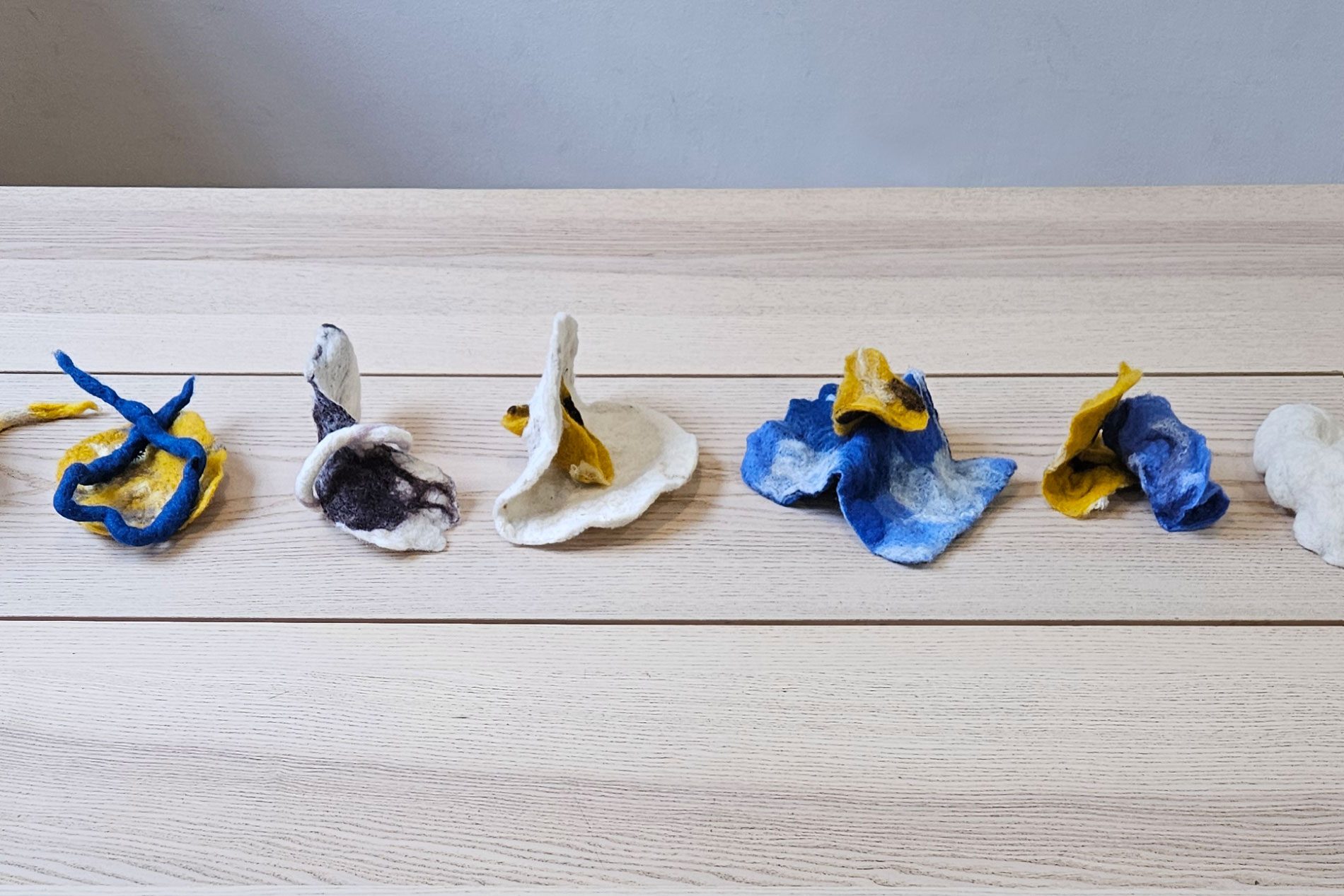 Several small felt sculptures with flexible wire armatures, in shades of blue, yellow and white. They are arranged in a line on a wooden table.
