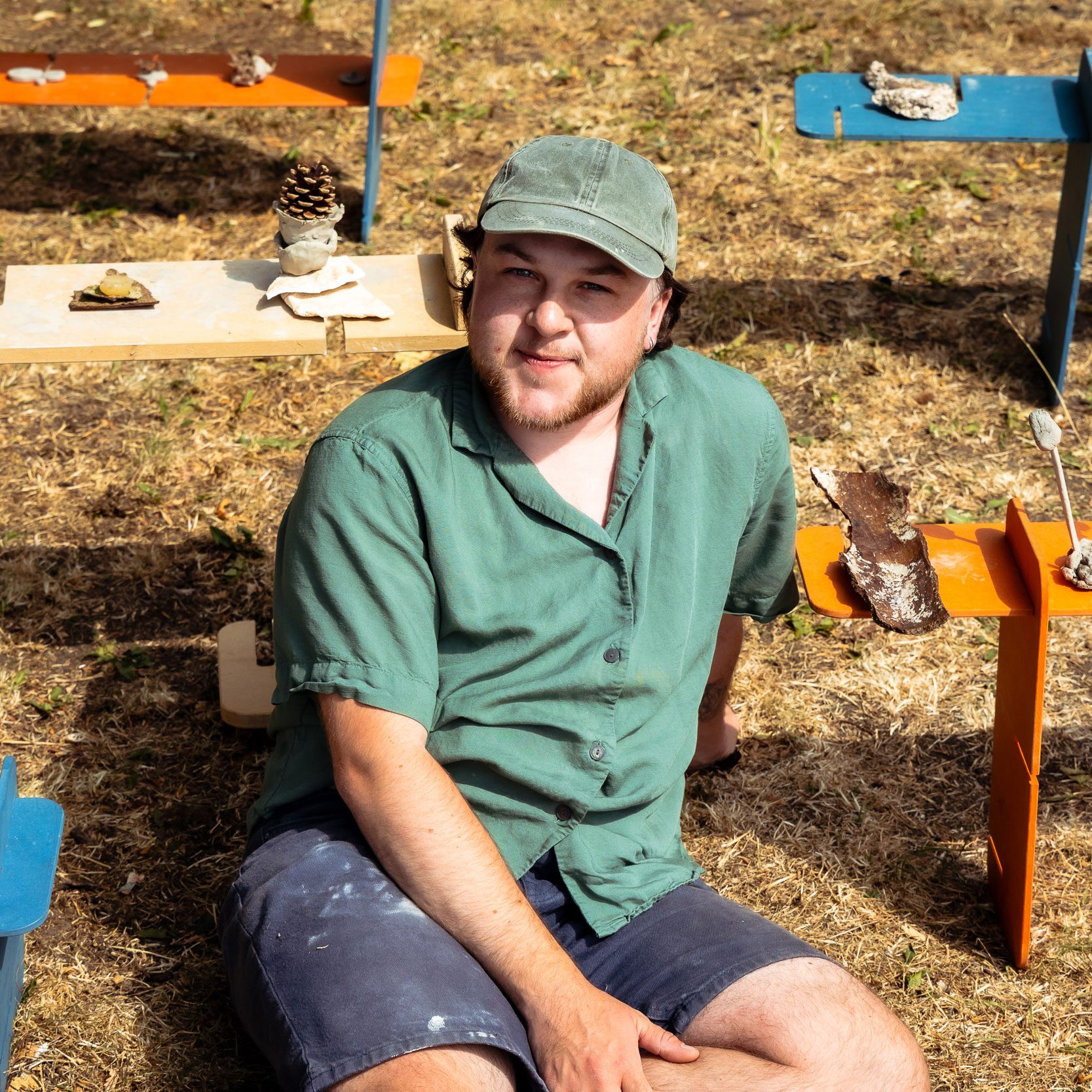 A man sitting on a grassy field, surrounded by several low benches supporting natural materials and community-made sculptures.