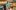 A man sitting on a grassy field, surrounded by several low benches supporting natural materials and community-made sculptures.